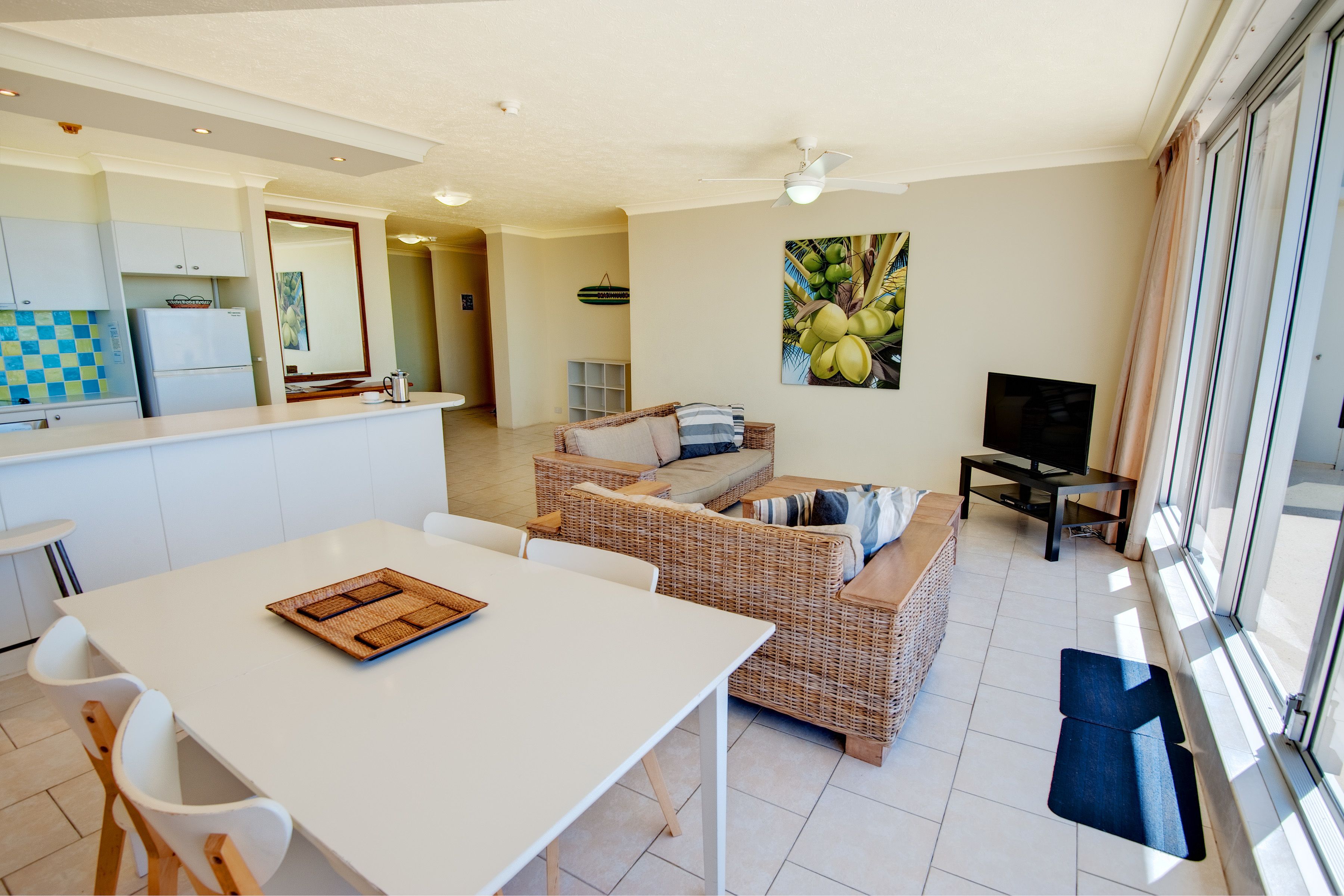 A modern kitchen with white cabinets, a dining table with chairs, a wicker sofa, and a TV.