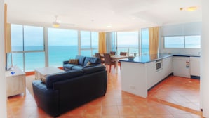 A spacious living area with a large sofa, dining table, and chairs, a kitchen with a central island, and large windows offering a view of the ocean.