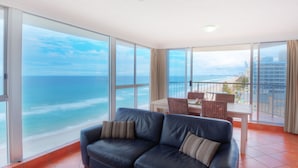 A modern living room with a large window overlooking the ocean, a dark blue leather sofa, and a dining area with a table and chairs.