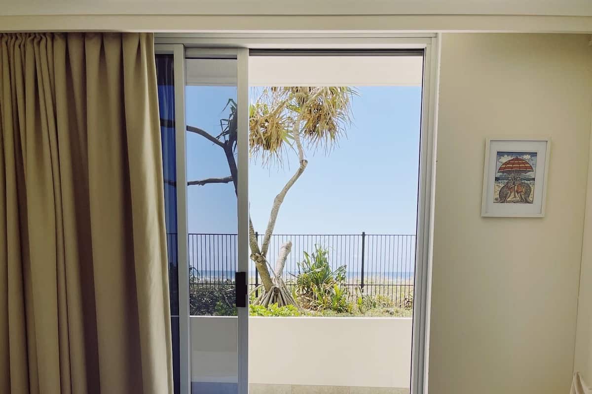 A hotel room with a bed, a chair, a balcony with a view of the beach and palm trees, and a framed picture on the wall.