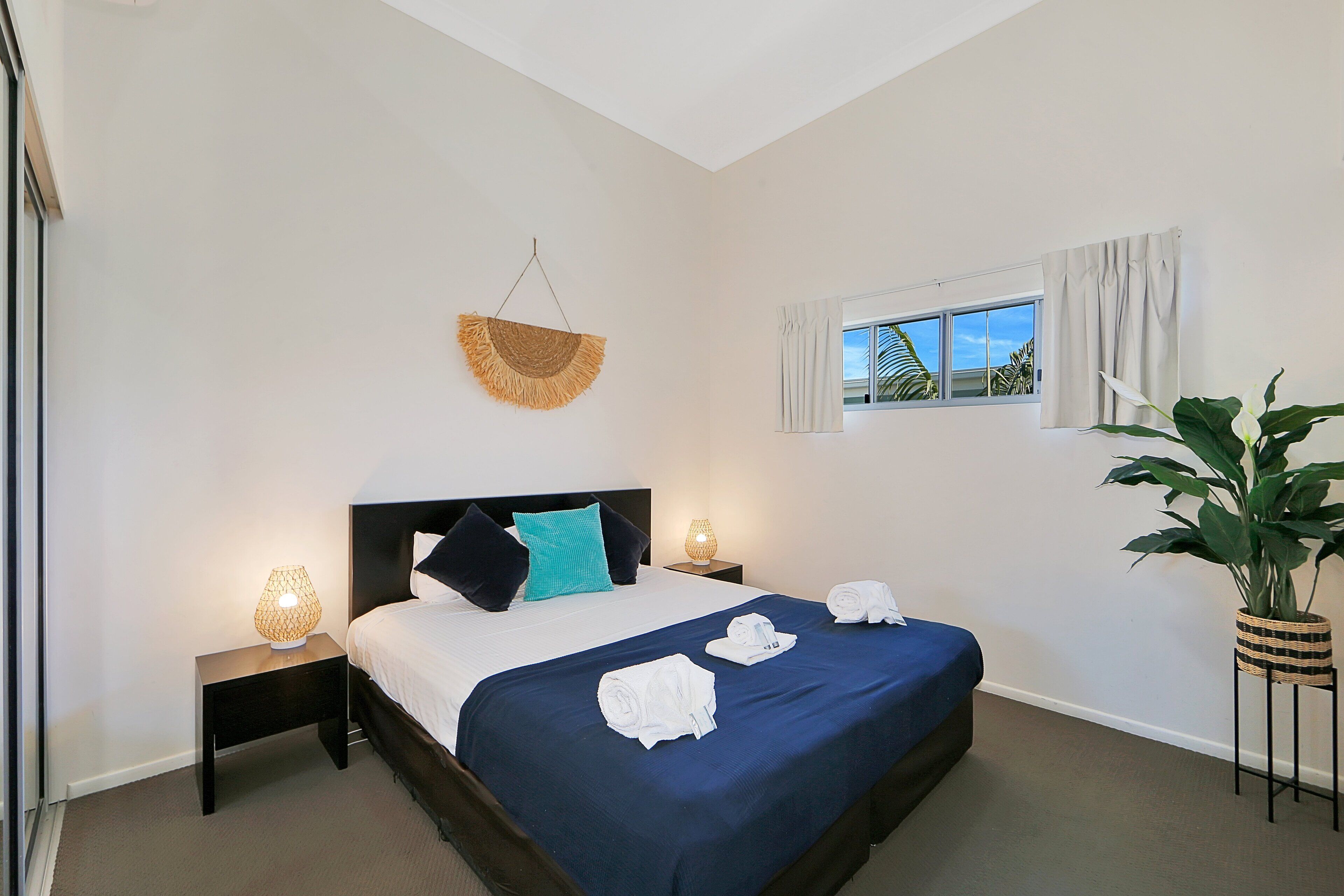 A neatly arranged hotel room with a large bed, two bedside tables with lamps, a window with curtains, and a potted plant.