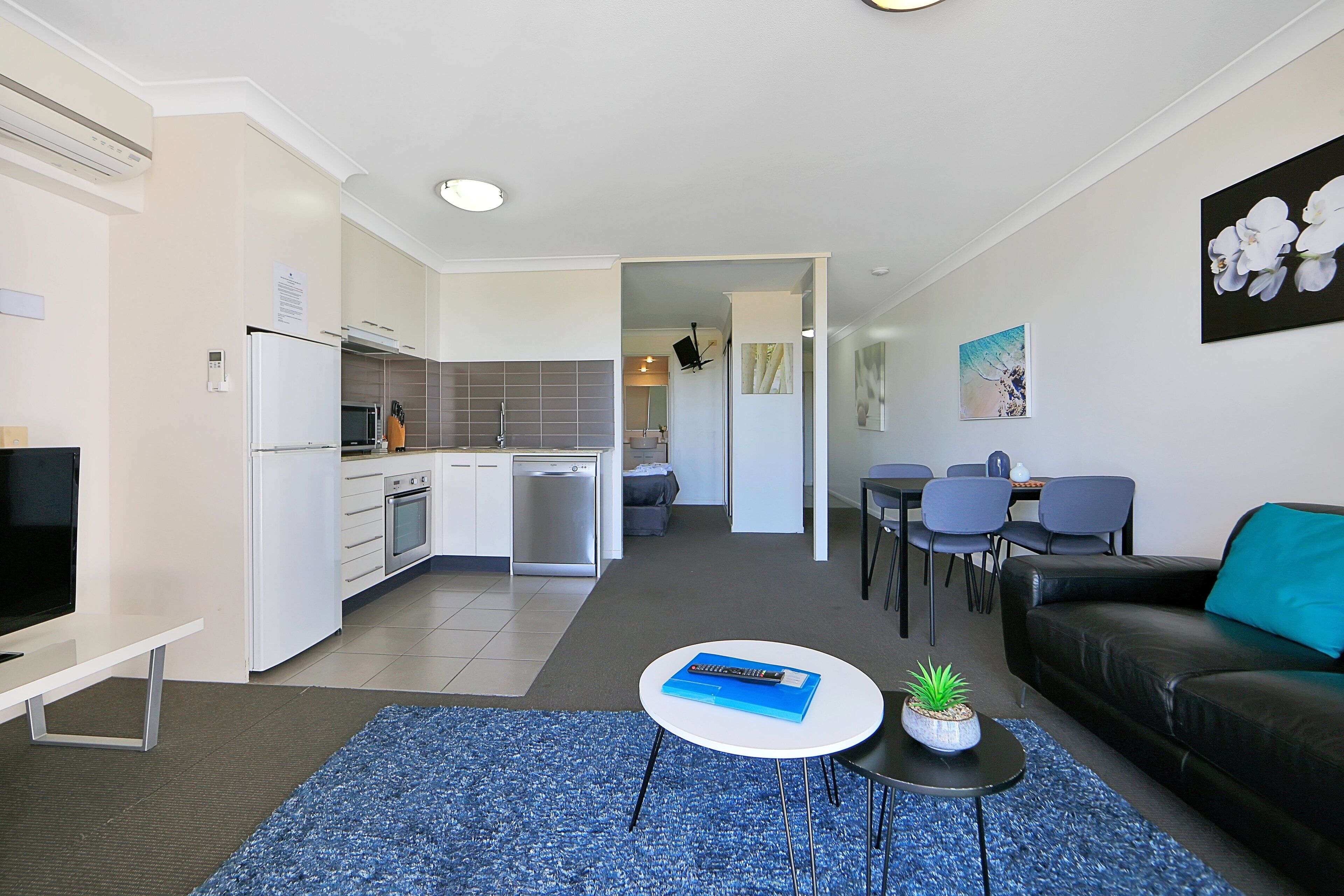 A modern living room with a sofa, a coffee table, and a TV. There is a kitchen in the background with a dishwasher and a microwave.