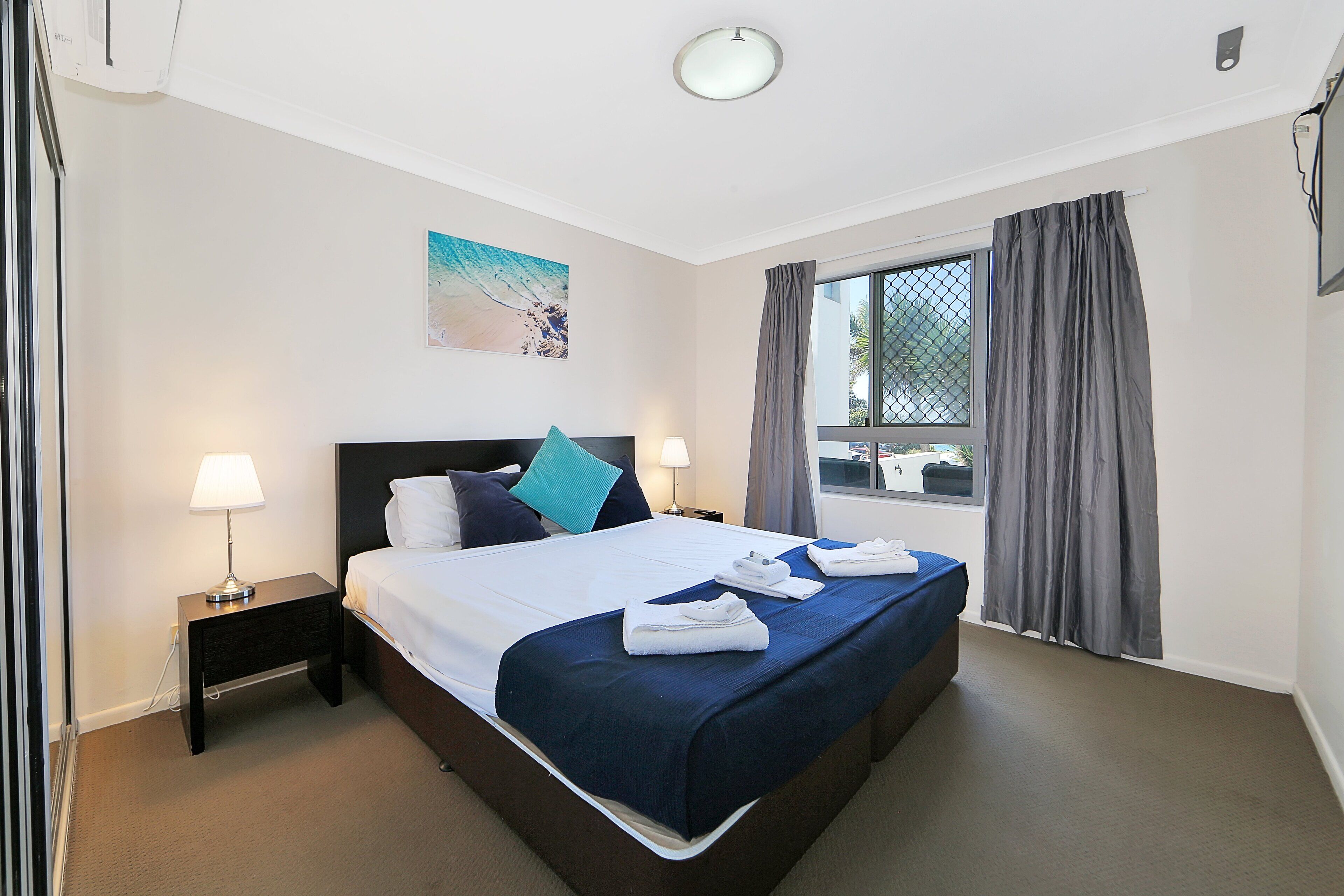 A neatly made bed with a blue bedspread, two pillows, and a small bedside table with a lamp.