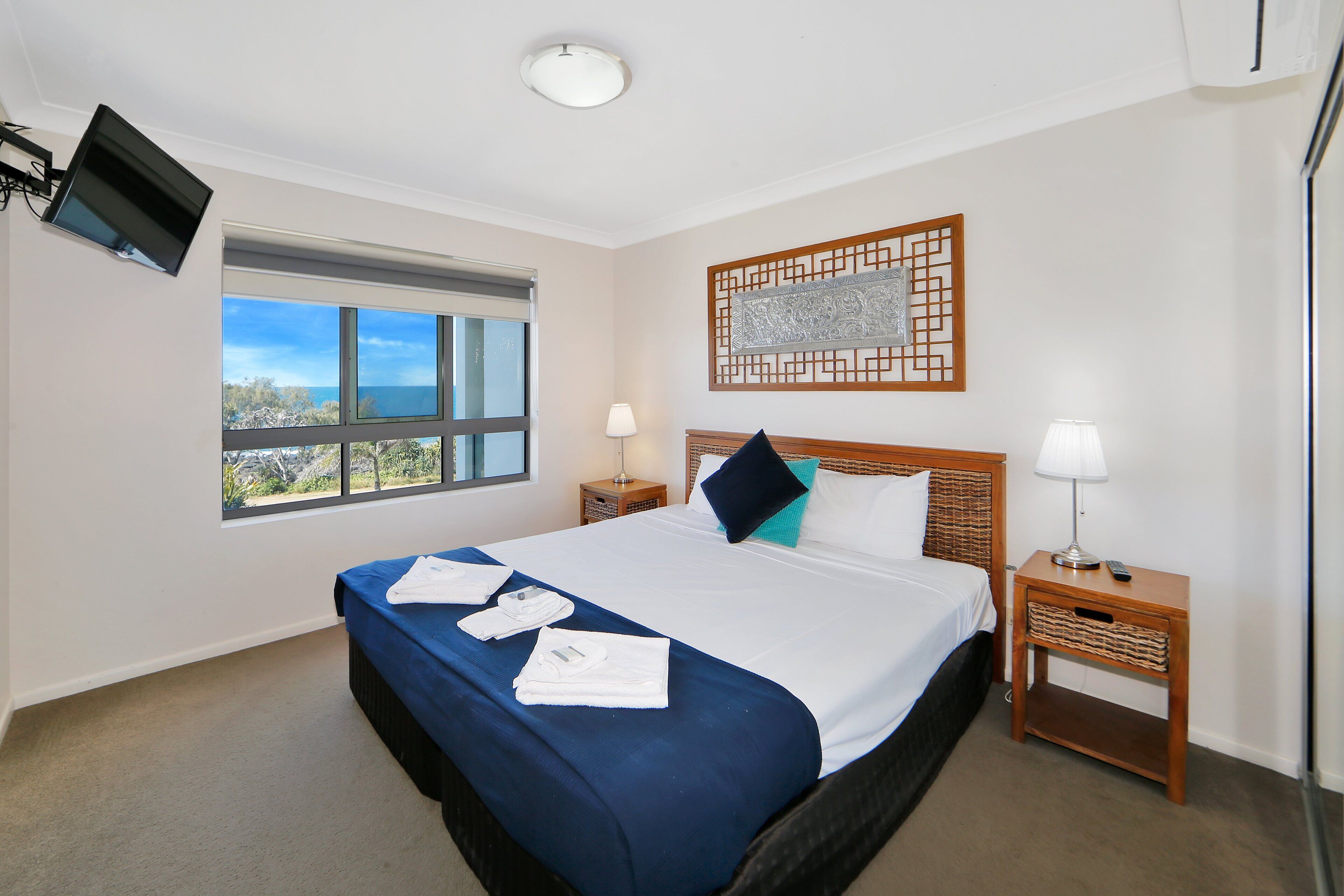 A hotel room with a large bed, two bedside tables, a TV mounted on the wall, a window with a view, and a decorative wall piece.