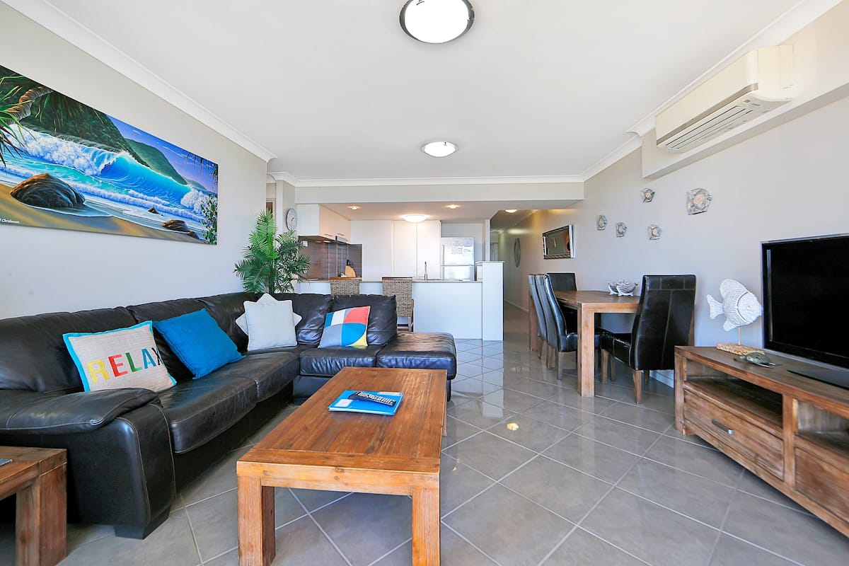 Uma sala de estar moderna com um sofá de couro preto, uma mesa de centro de madeira e uma televisão de tela plana. Há uma área de jantar com mesa e cadeiras, e uma cozinha ao fundo.