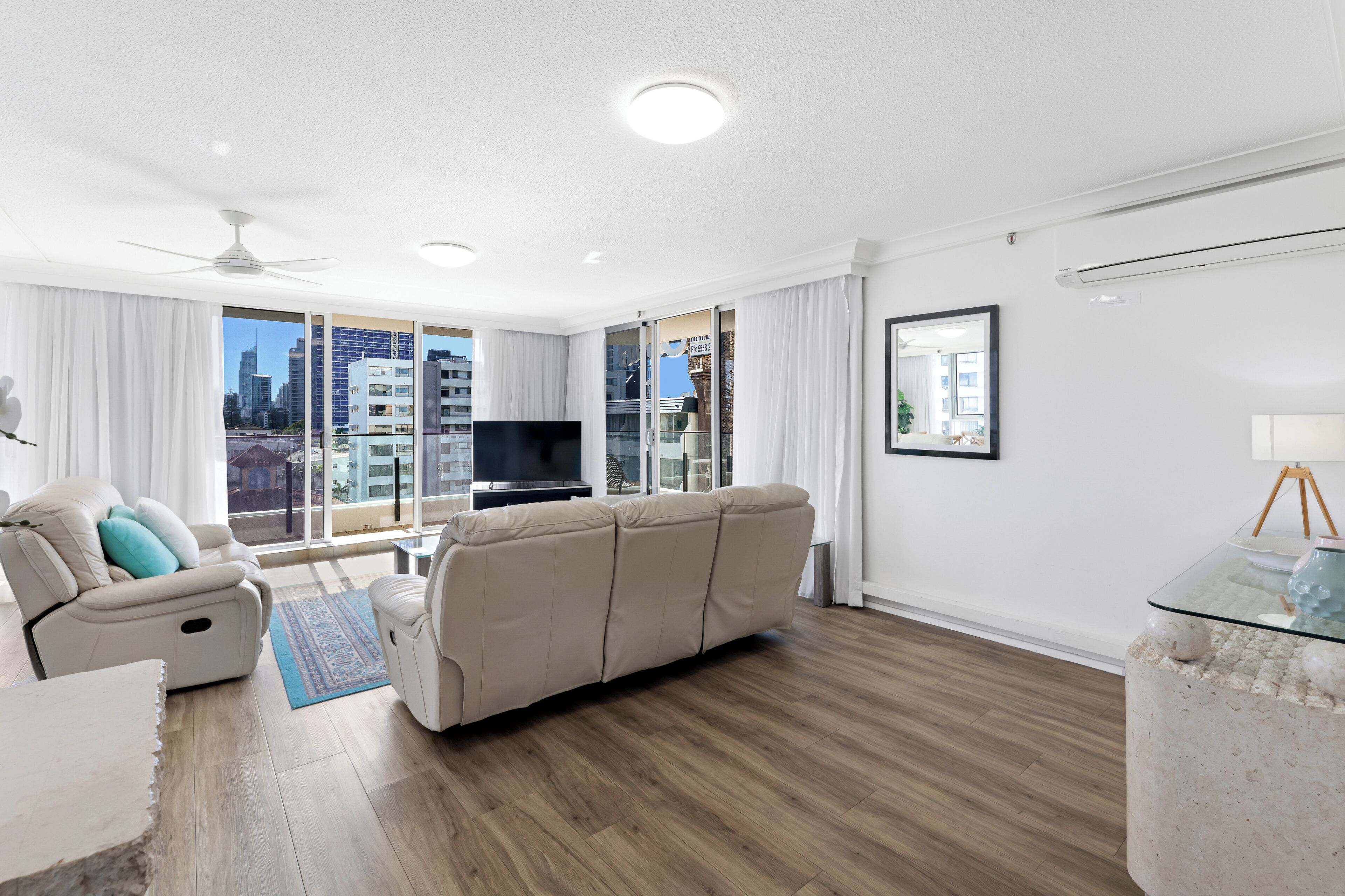A modern living room with a beige sofa, a white armchair, a glass coffee table, and a flat-screen TV.
