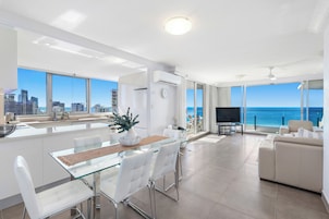 A modern living room with a glass dining table, white chairs, a beige sofa, and a TV.
