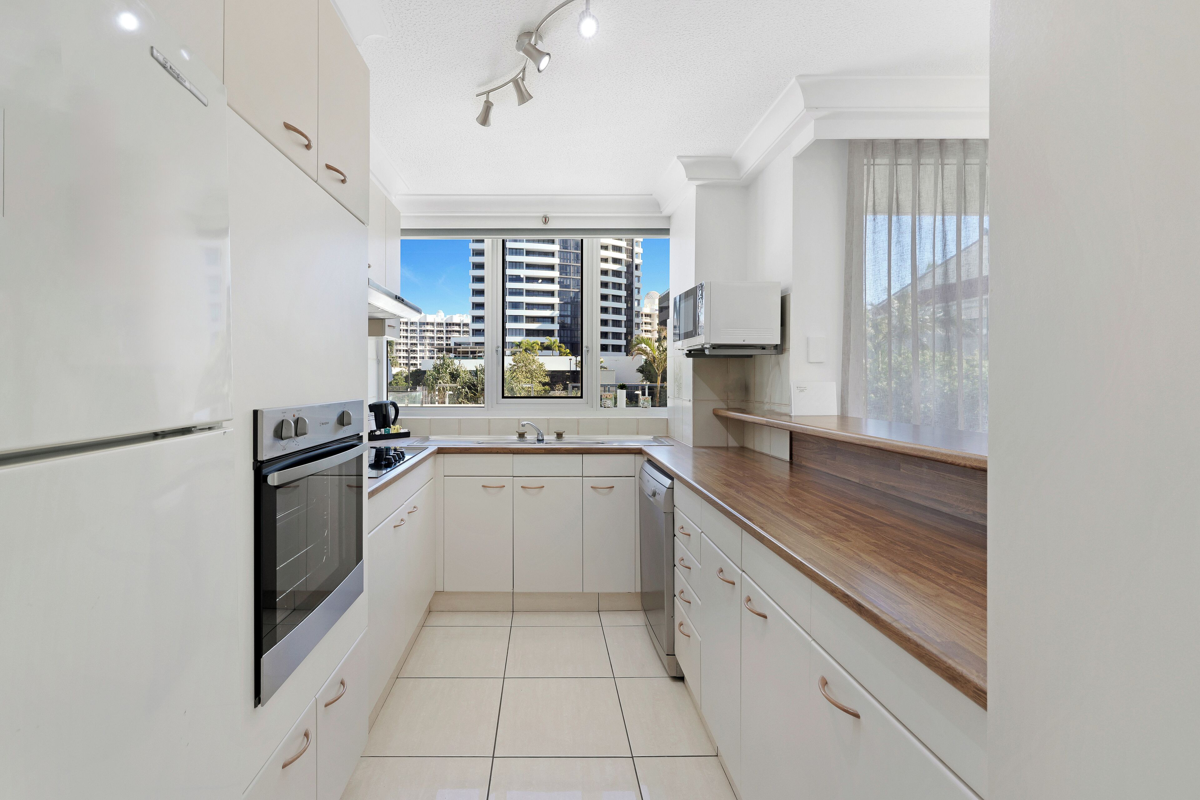 A modern kitchen with white cabinets, a wooden countertop, and a large window offering a view of tall buildings.