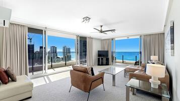 A spacious living room with a glass coffee table, a sofa, and a TV. Large windows offer a view of the city and ocean.