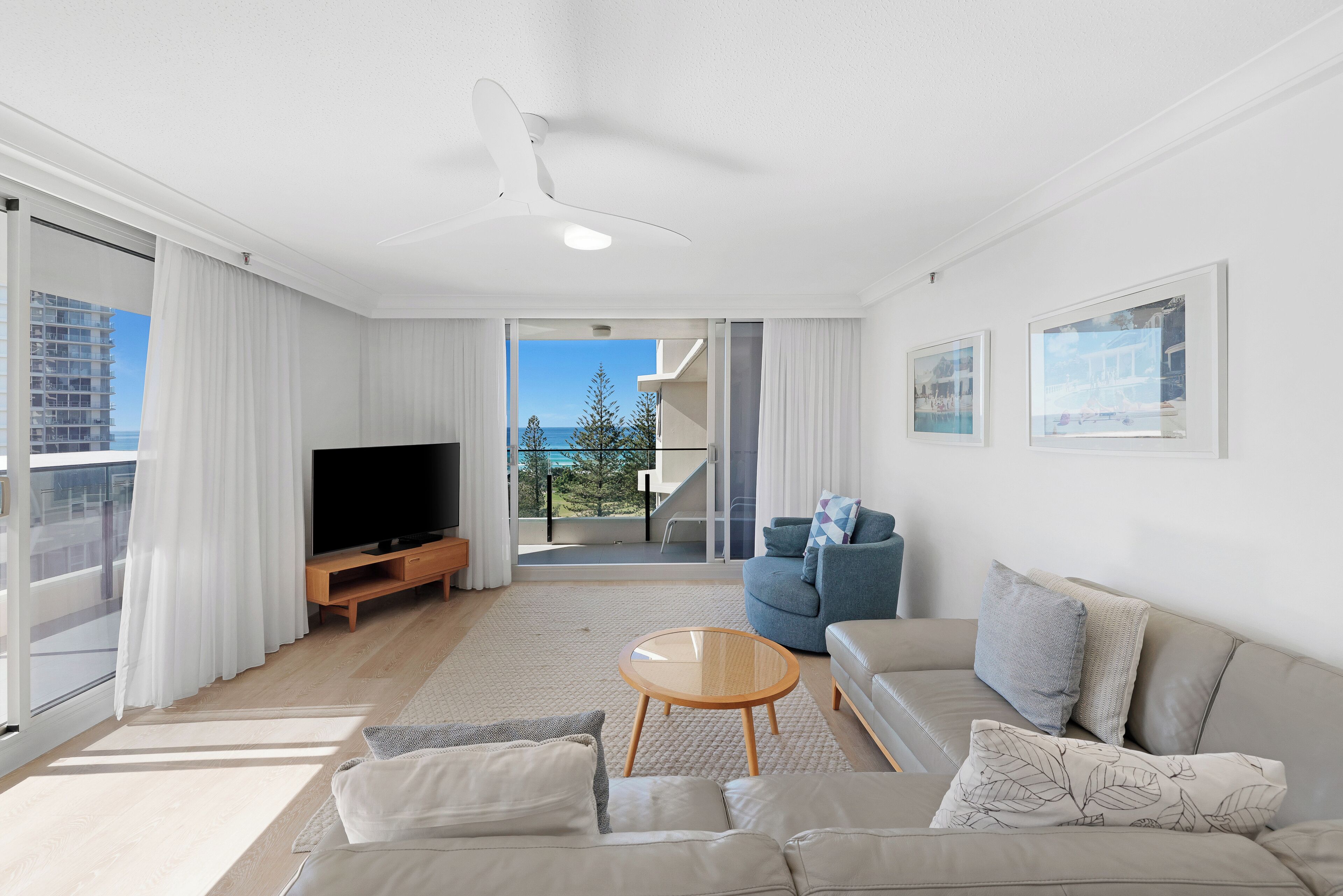 A modern living room with a flat-screen TV, a sofa, a coffee table, and a balcony with a view of the sea.
