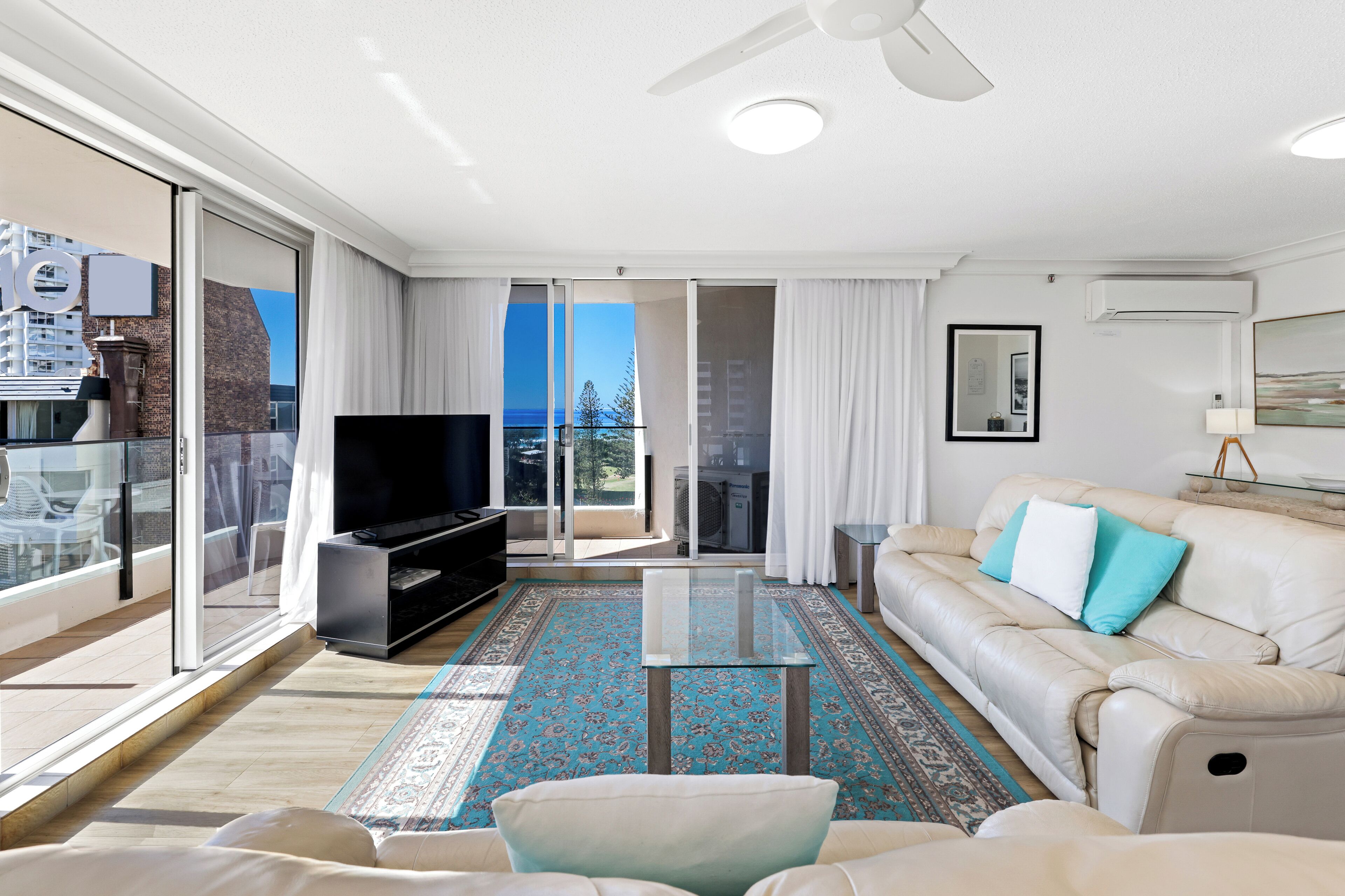 Een moderne woonkamer met een bank, een glazen salontafel, een flatscreen televisie en een grote schuifpui naar een balkon.