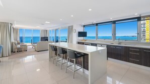 A modern kitchen with a central island, bar stools, and a large window overlooking the sea. - ULTIQA Beach Haven At Broadbeach (Broadbeach)
