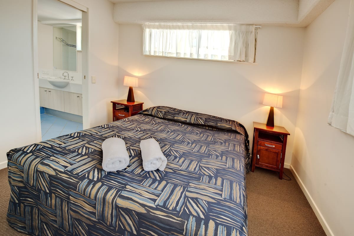 A bedroom with a bed, two bedside tables with lamps, a window with curtains, and a bathroom visible through an open door.