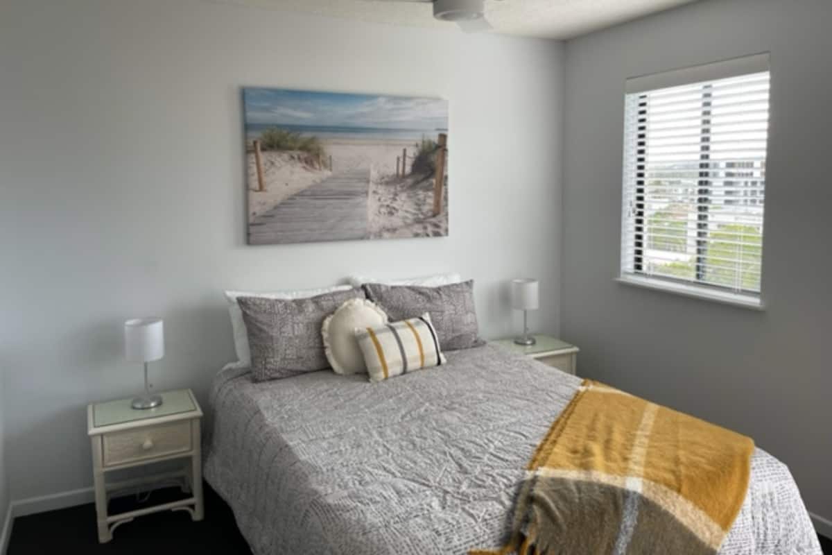 A bedroom with a bed, a ceiling fan, a window with blinds, a nightstand, and a wall-mounted picture.