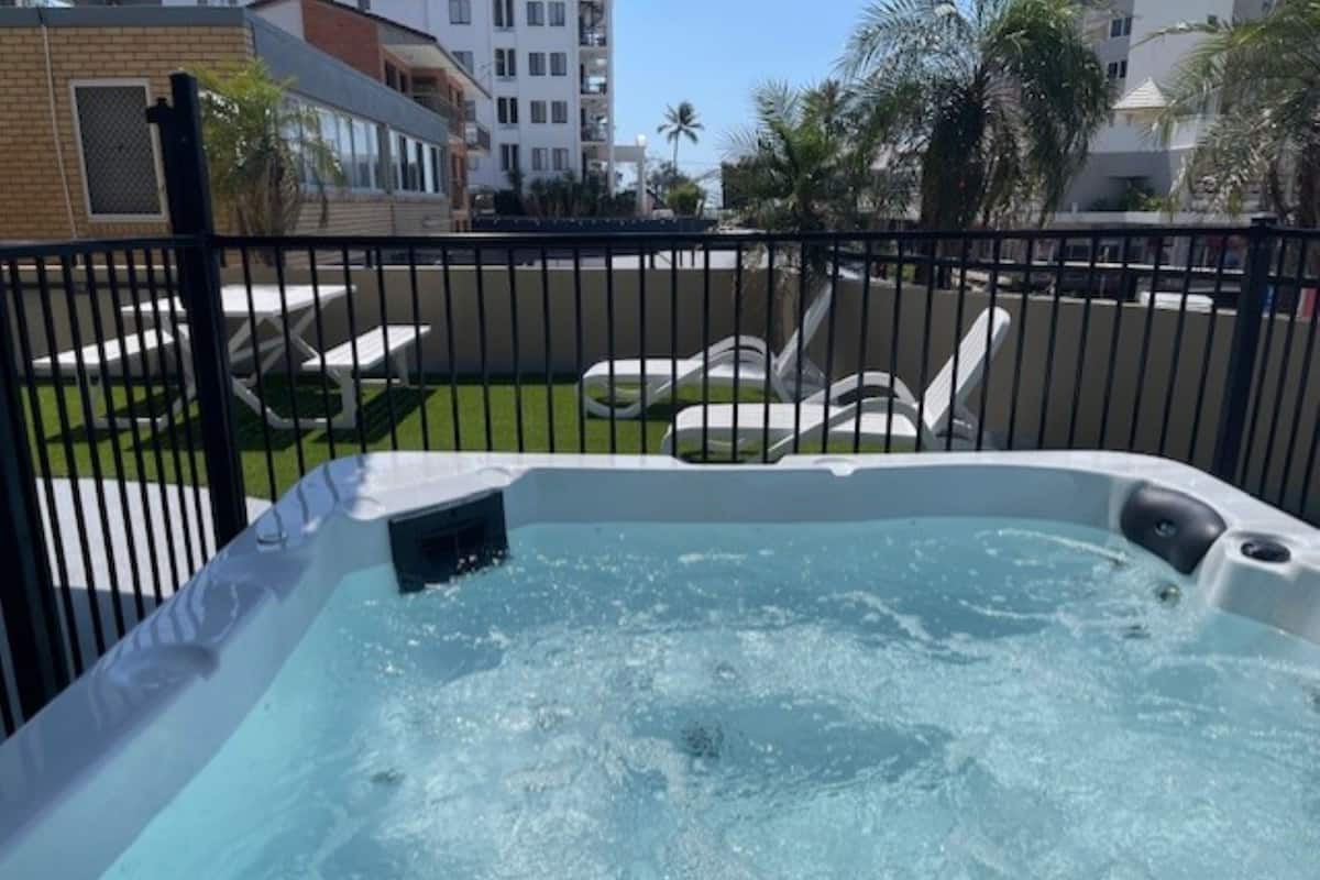 A hot tub surrounded by a black fence, with a grassy area and lounge chairs in the background.