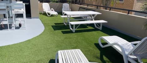 A rooftop terrace with artificial grass, white lounge chairs, and a picnic table.