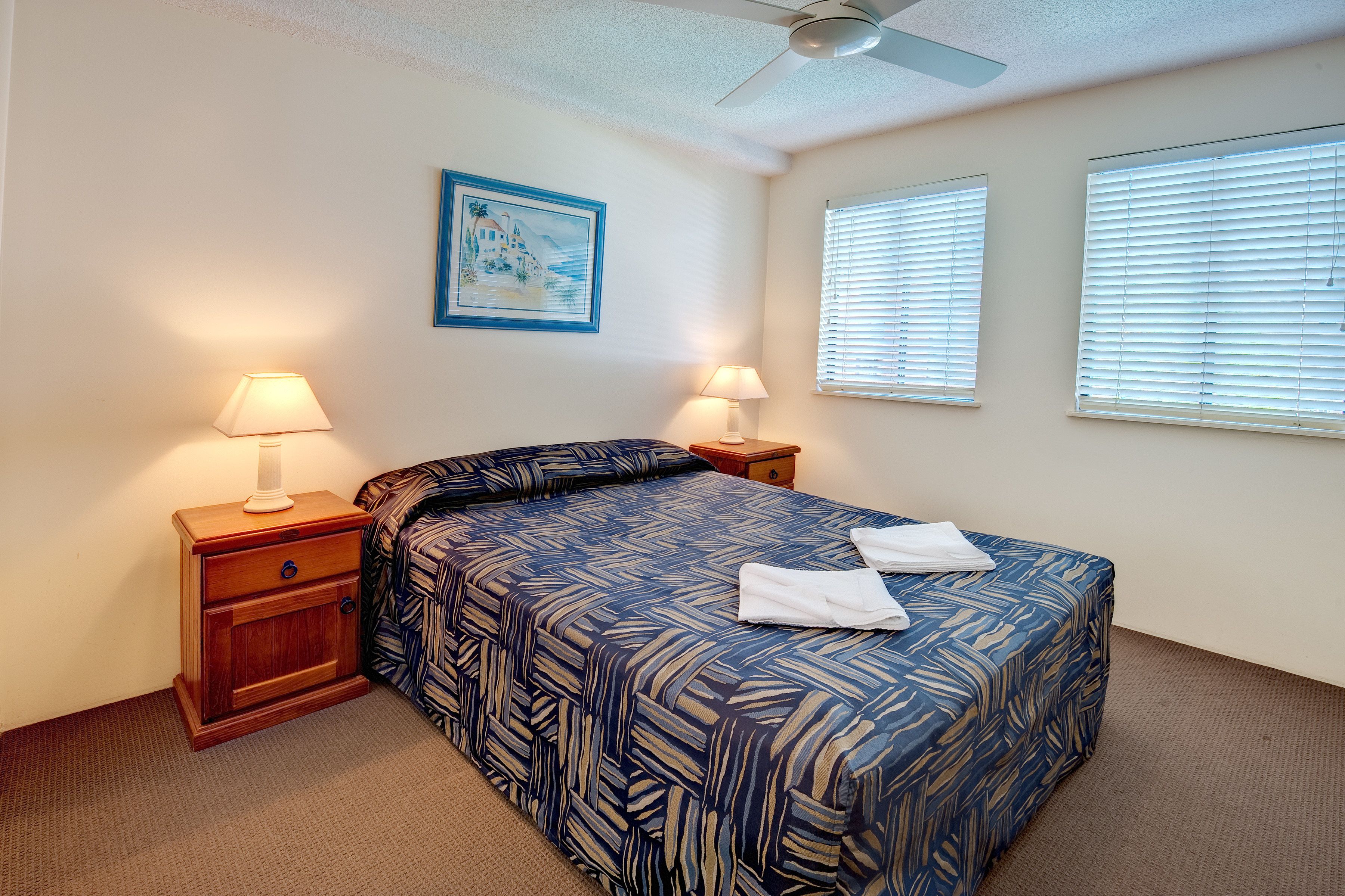 A neatly made bed with a patterned bedspread, two bedside lamps, a nightstand, a ceiling fan, and a framed picture on the wall.