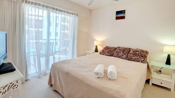 Habitación de hotel con cama, dos lámparas de noche, televisor sobre una mesita auxiliar y vistas al exterior a través de amplios ventanales.