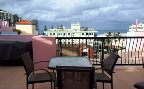 A balcony with a table and chairs, overlooking a building with a view of the sea.