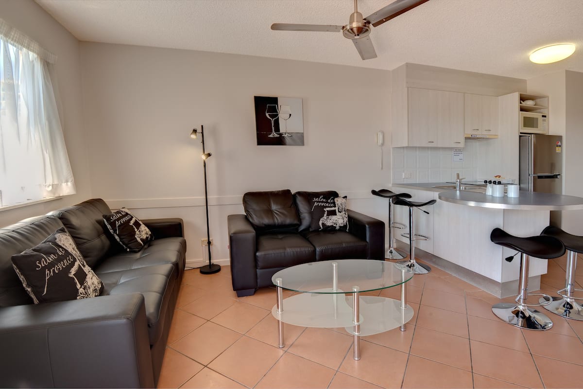 A modern living room with a glass coffee table, a black leather sofa, and a black leather couch. There is a kitchen area with white cabinets and a stainless steel refrigerator.