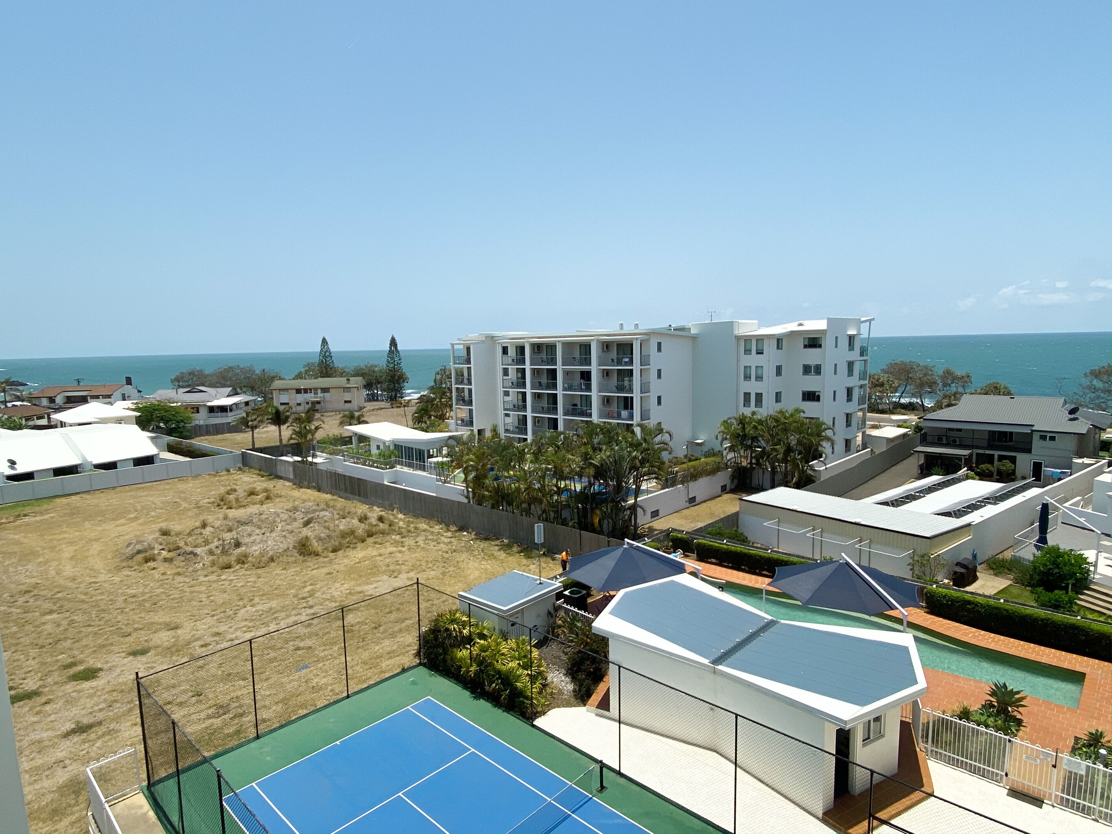 A residential area with a tennis court, a fenced yard, and a building with multiple balconies overlooking the ocean.
