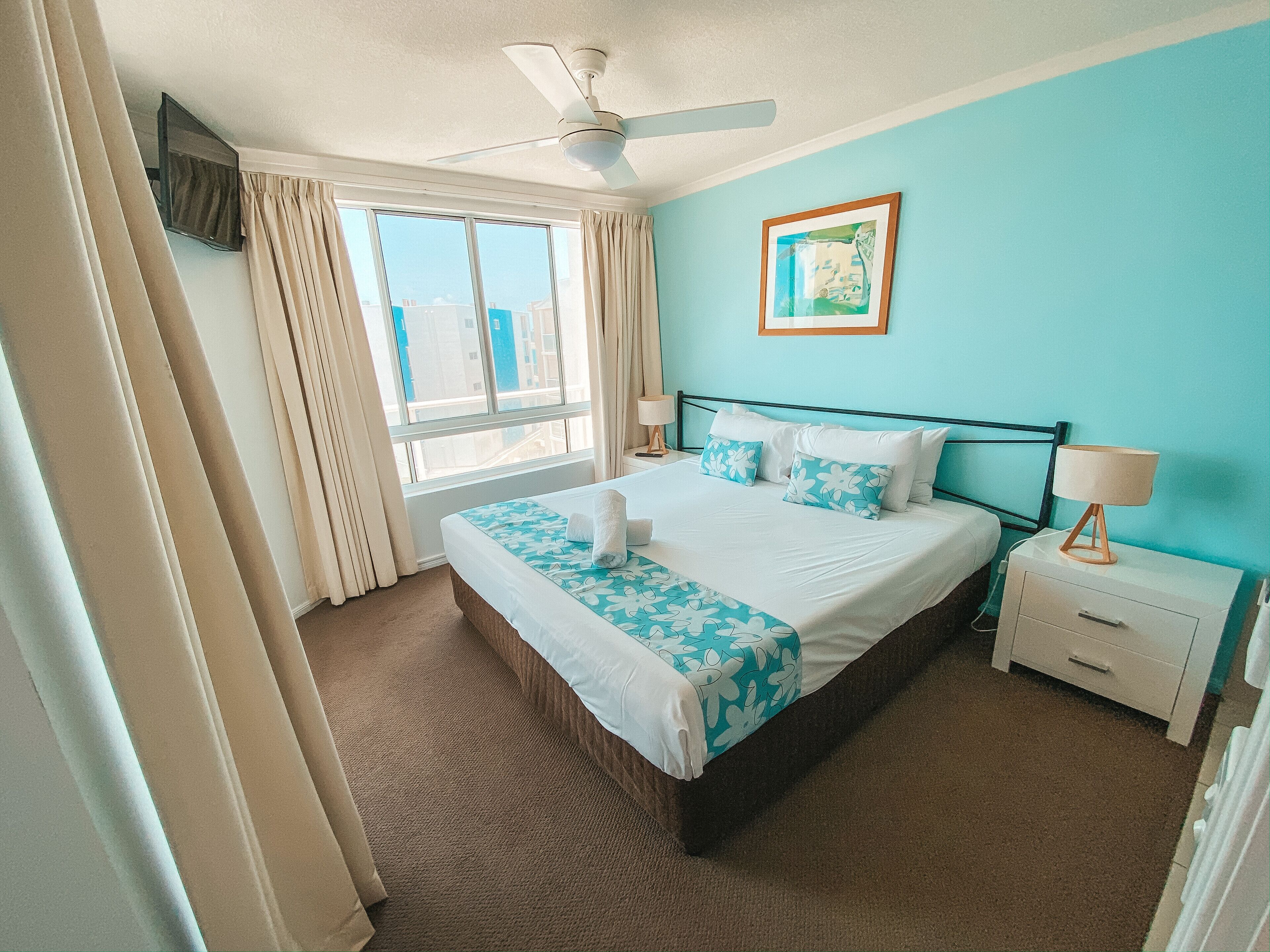 Une chambre d’hôtel avec un lit, une table de chevet, un ventilateur de plafond, une fenêtre avec des rideaux et un tableau au mur.
