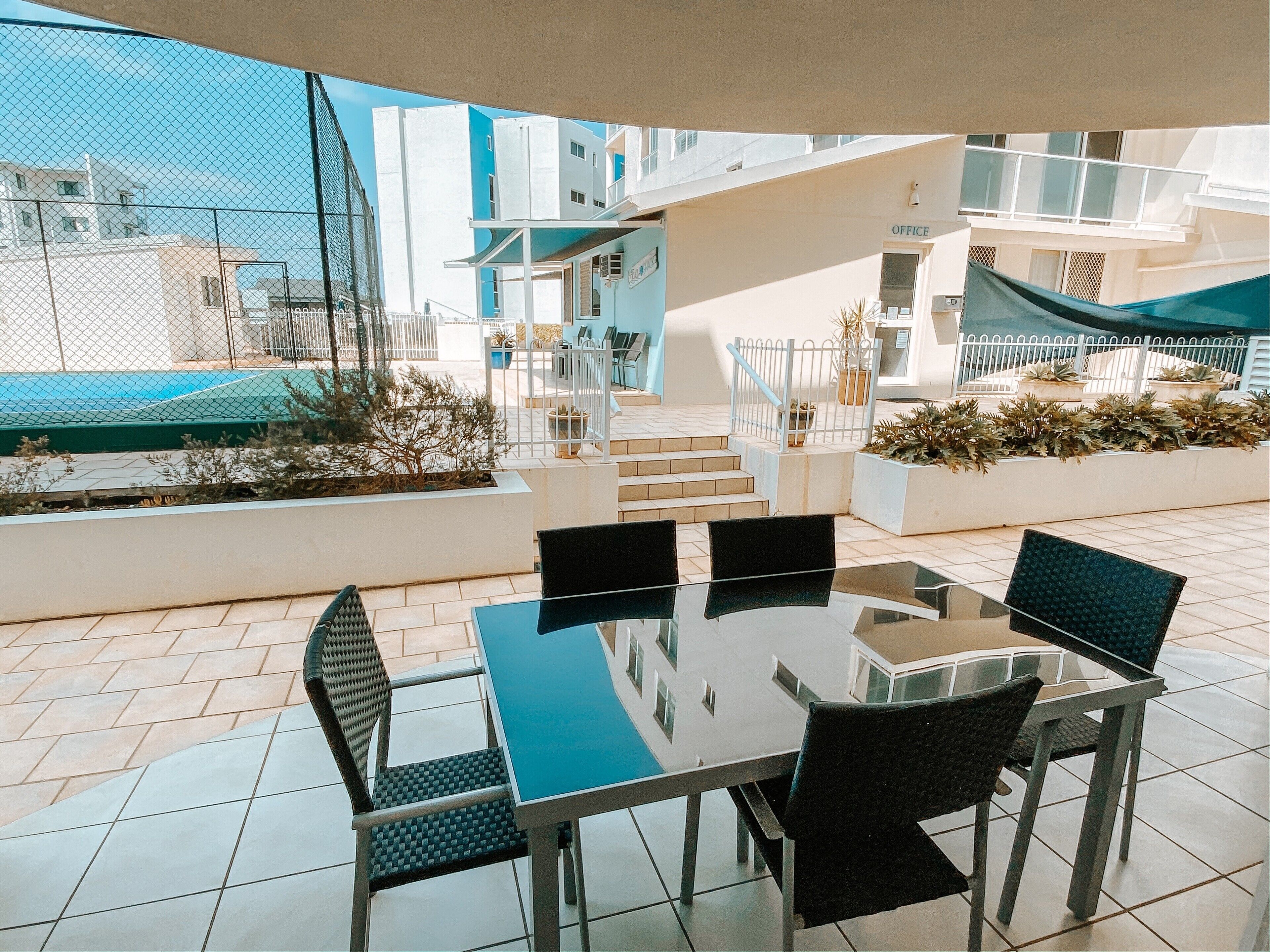 An outdoor patio with a glass table and four chairs, surrounded by a white fence and potted plants.