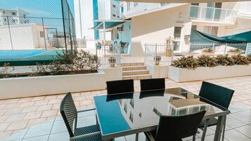 Un patio al aire libre con una mesa de vidrio y cuatro sillas, rodeado por una valla blanca y plantas en macetas.