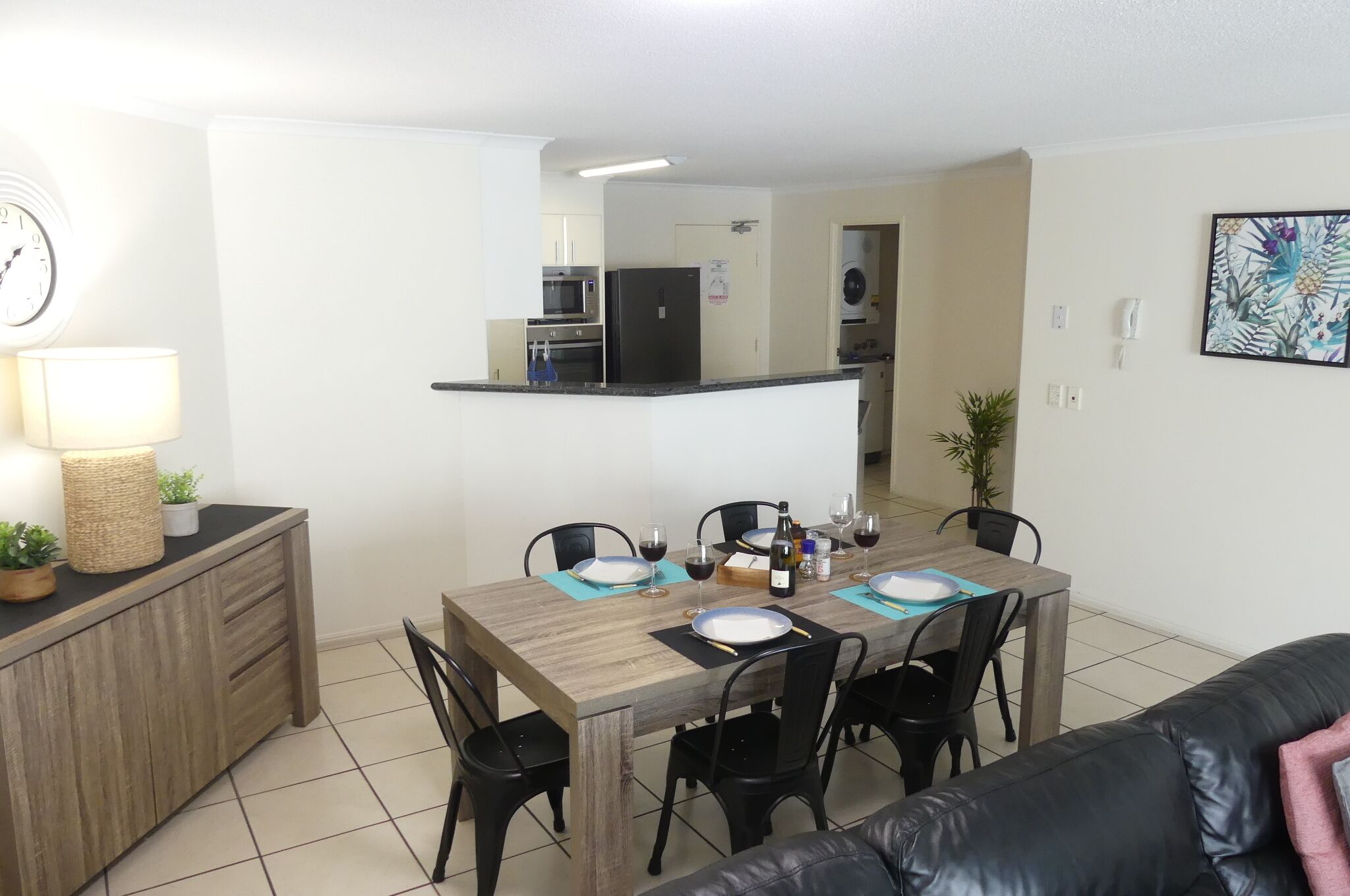 A dining area with a wooden table set for six, black chairs, and a kitchen in the background.