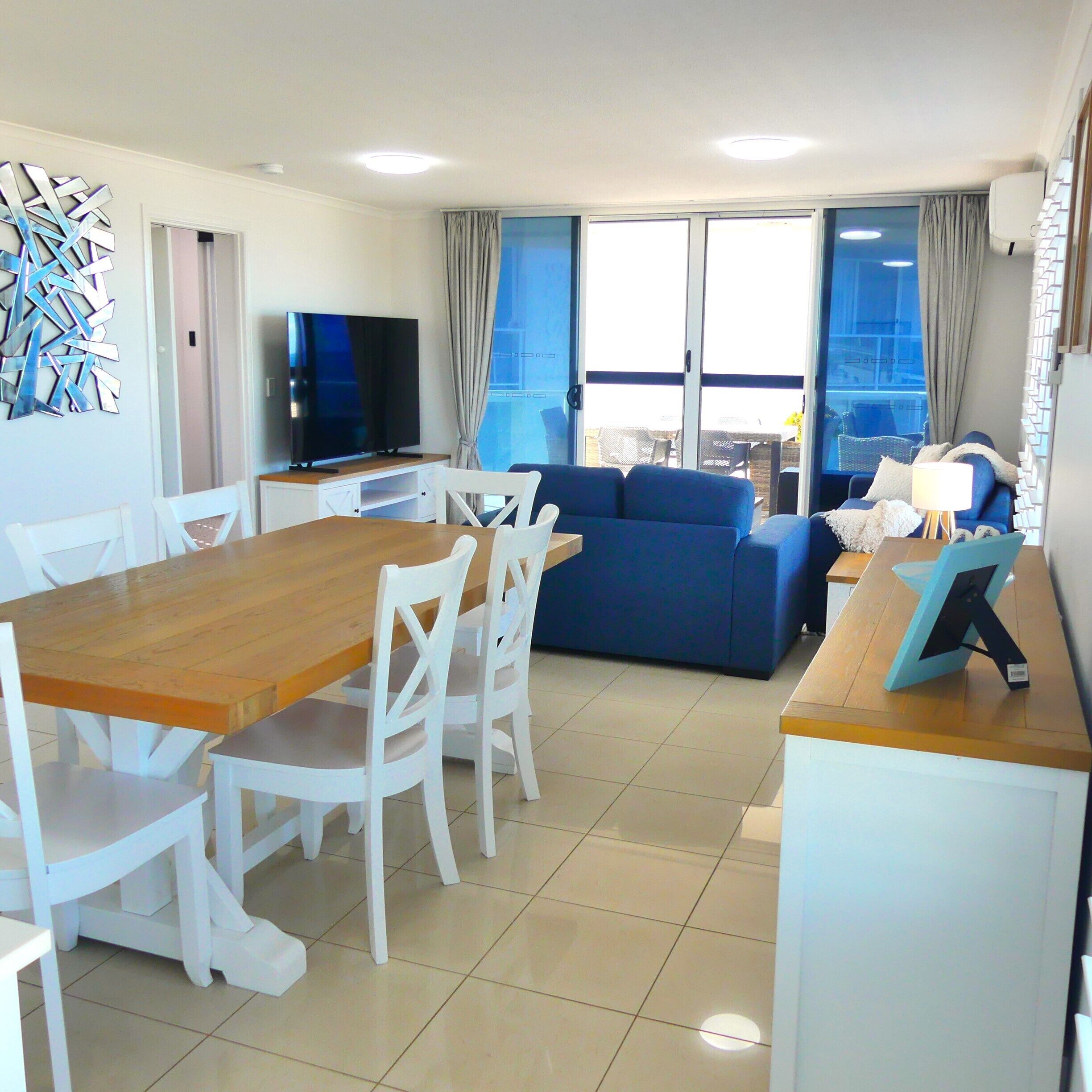 A modern living room with a wooden dining table, white chairs, a blue sofa, and a flat-screen TV.