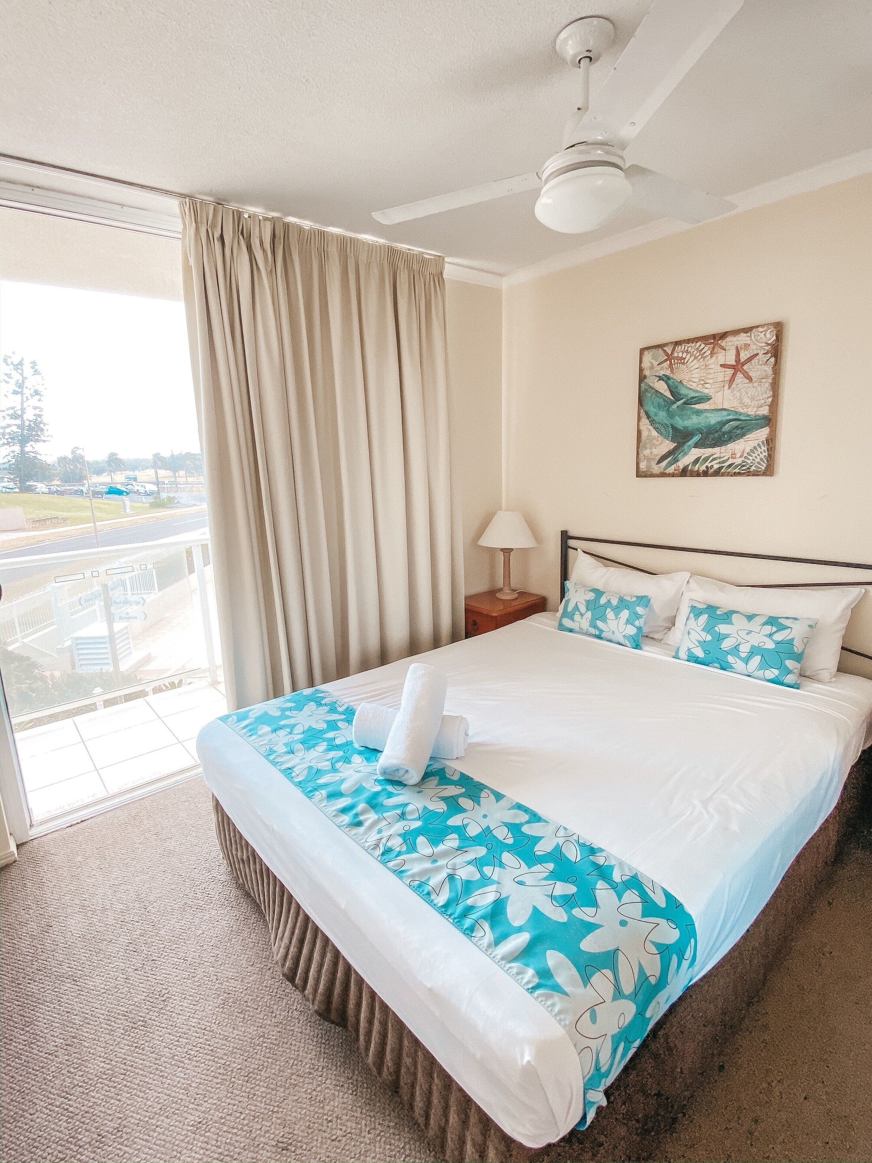 A hotel room with a bed, a nightstand, a lamp, a ceiling fan, a window with curtains, and a painting on the wall.