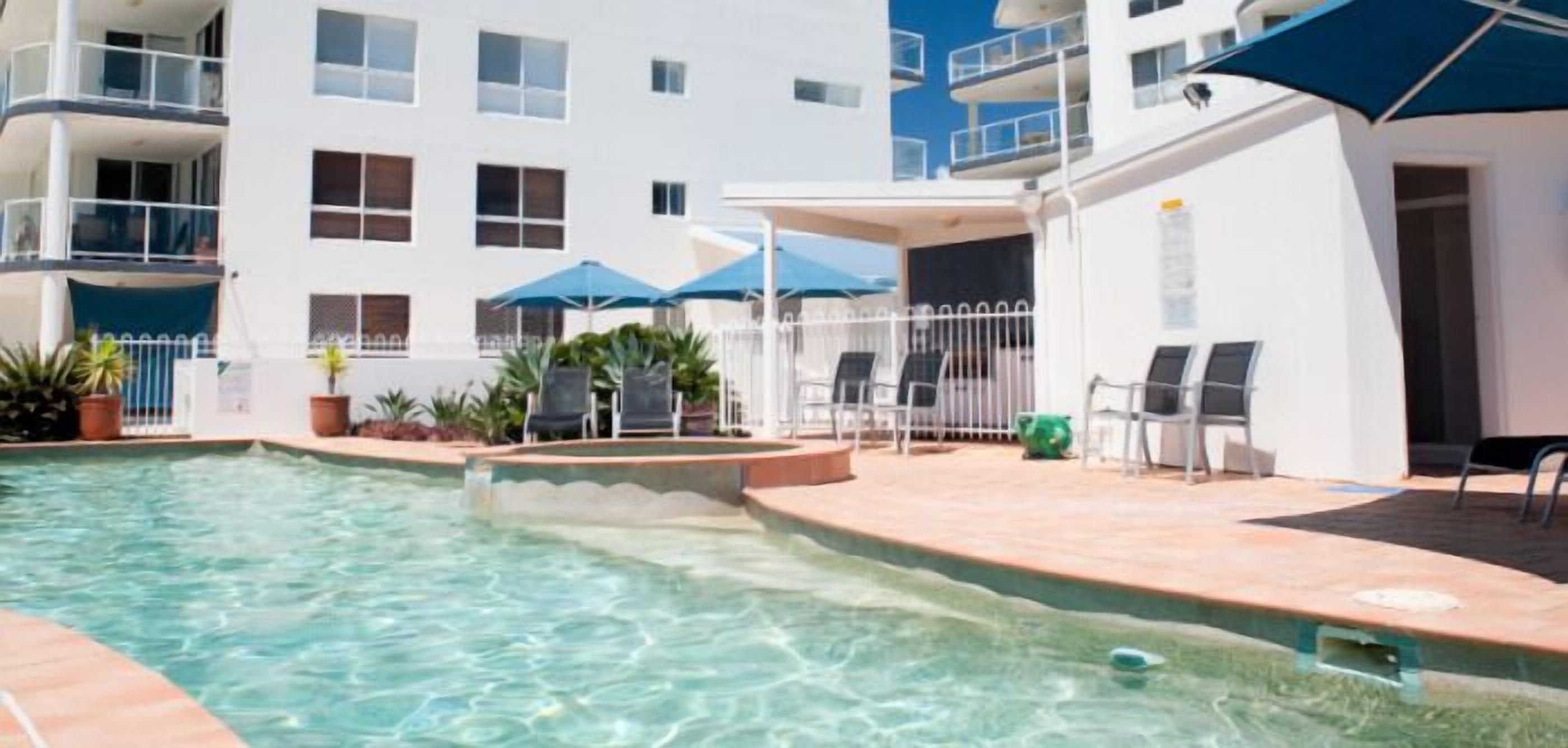 A swimming pool area with lounge chairs and umbrellas, surrounded by a building with balconies and windows.