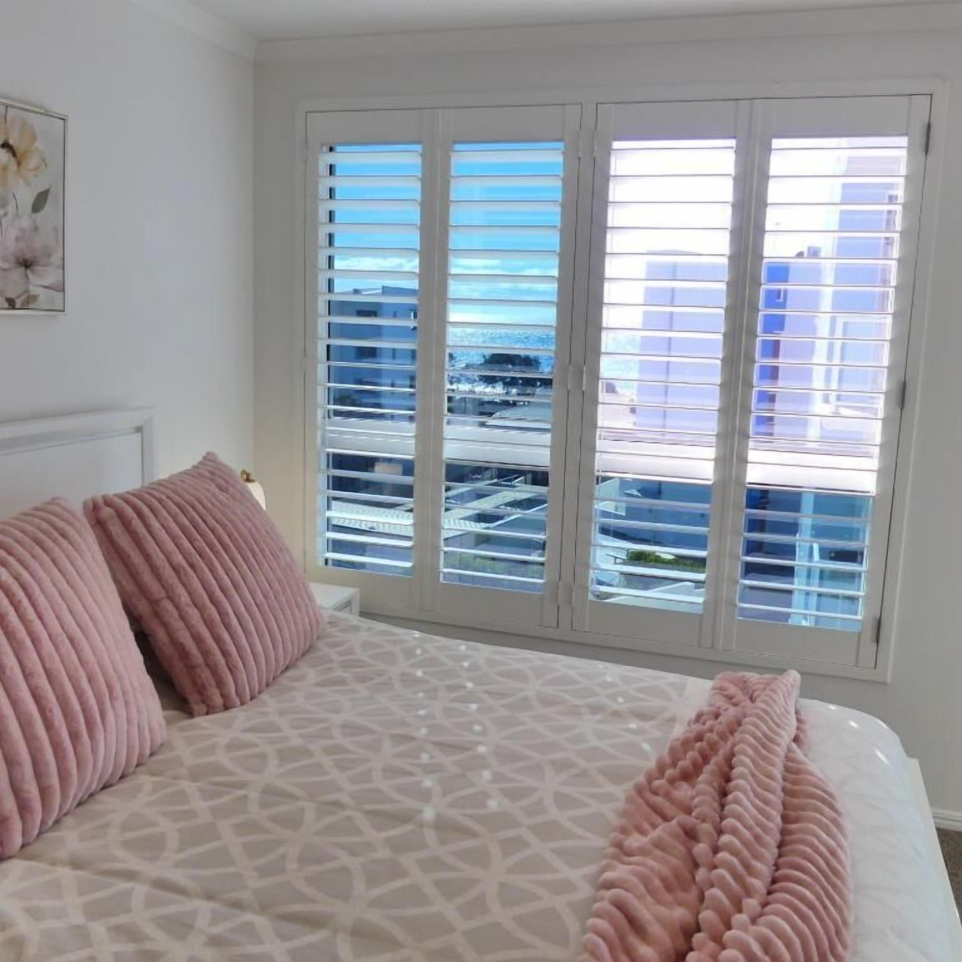A bedroom with a patterned bedspread, two pink pillows, and a view of buildings through large windows.