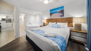 A modern hotel room with a large bed, a wooden headboard, a ceiling fan, a painting of a beach, and a view of the bathroom and kitchen.