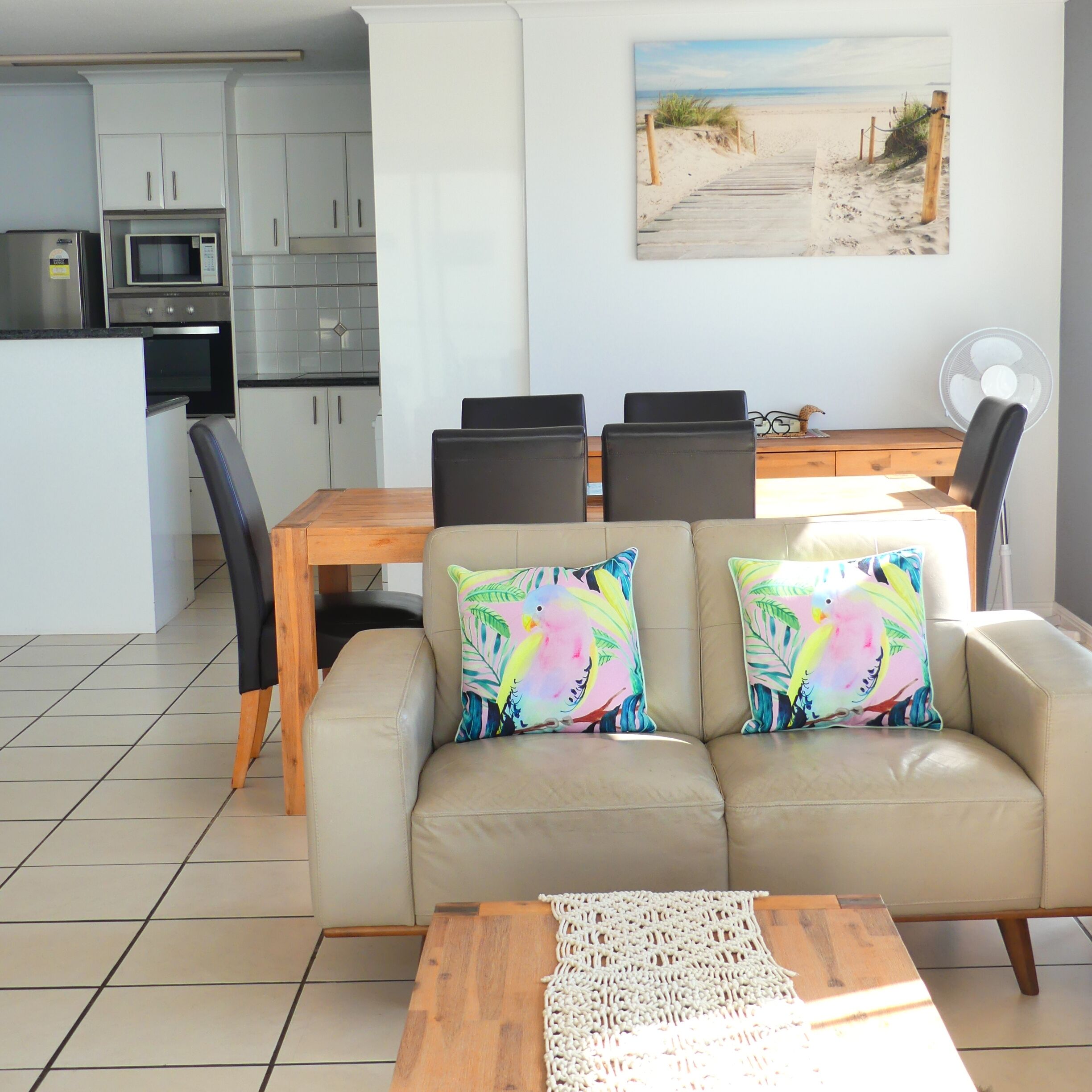 A living room with a beige sofa, two colorful pillows, a wooden coffee table, and a macrame table runner.