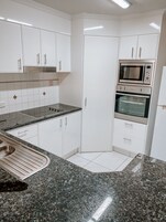Una cocina moderna con armarios blancos, un lavamanos de acero inoxidable, encimeras de granito y electrodomésticos integrados, incluyendo un microondas y un horno.