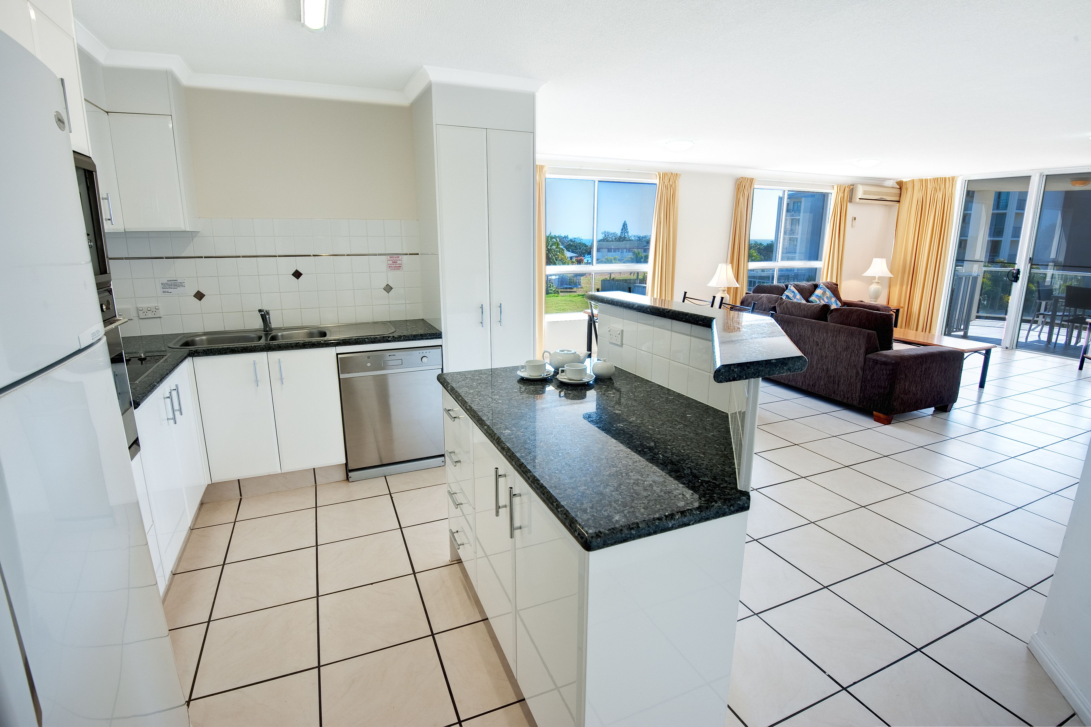 Cozinha moderna com armários brancos e bancada preta, área de jantar com sofá e mesa, e varanda com vista.