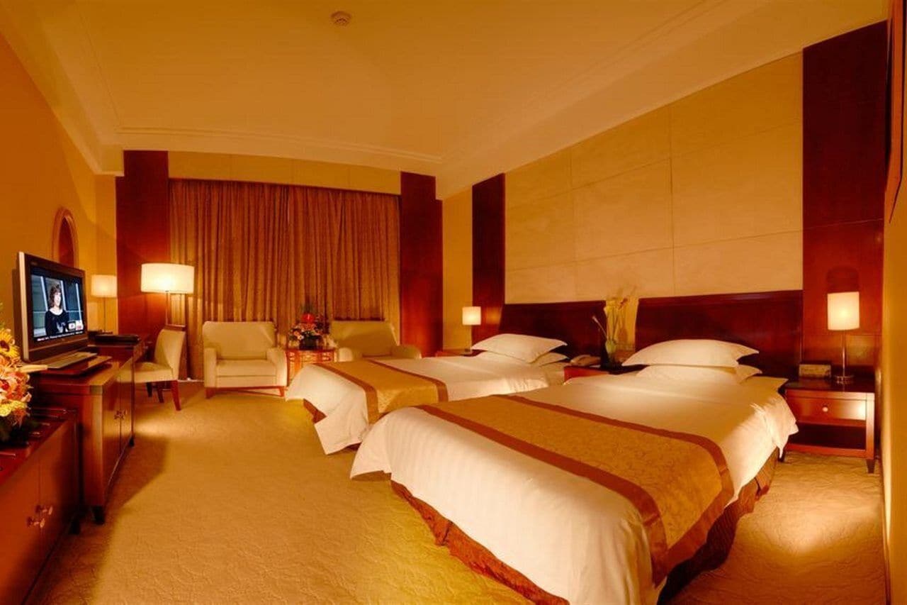A hotel room with three beds, a TV, a desk with a lamp, and a large window with curtains.