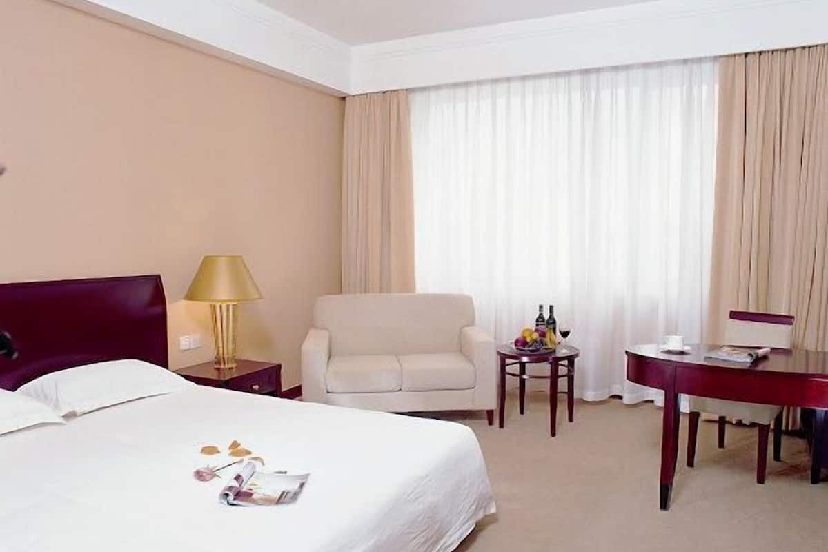 A hotel room with a bed, a desk, a chair, a lamp, and a table with a fruit bowl and bottles.
