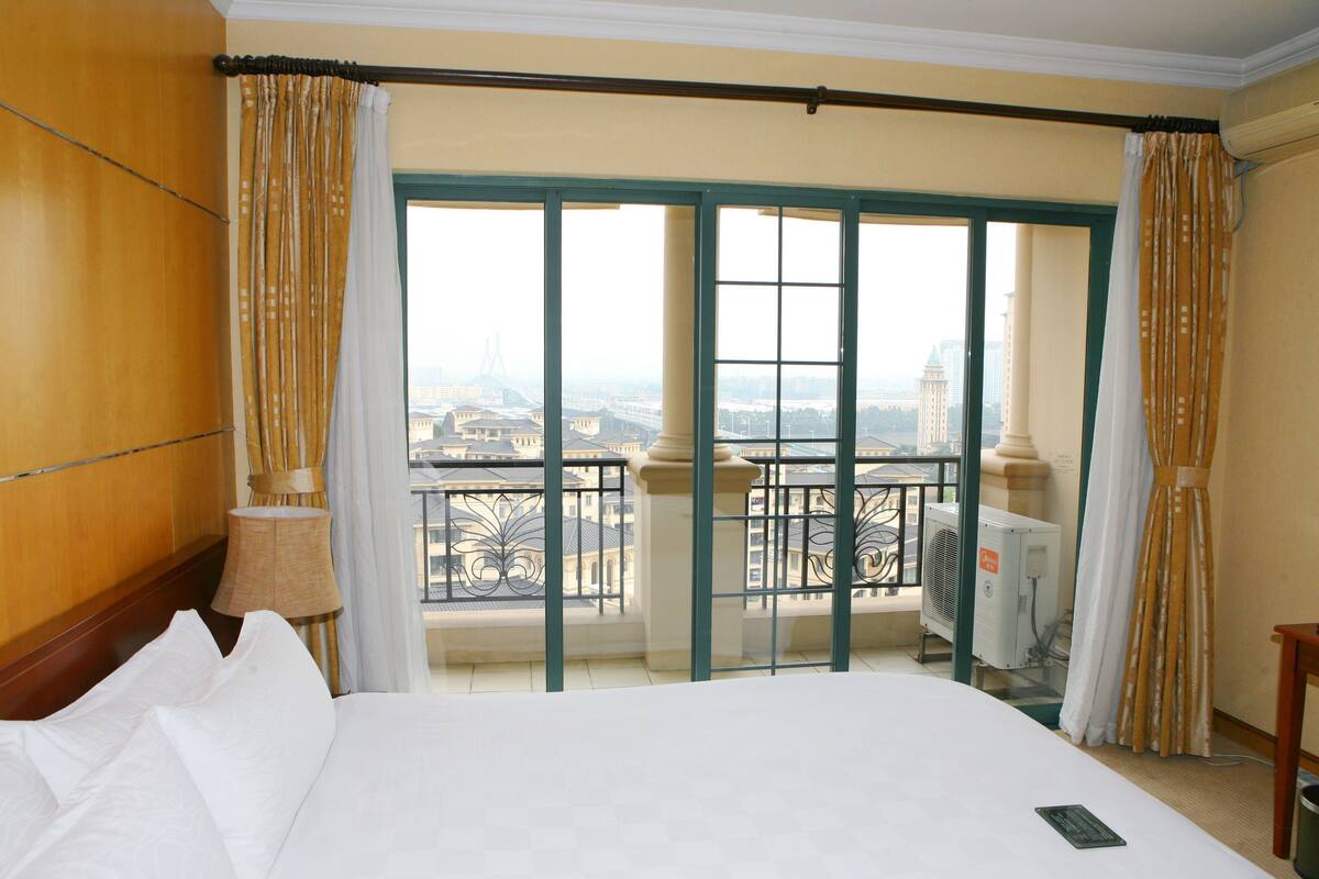 A hotel room with a large bed, a view of the city through a balcony, and a window with curtains.