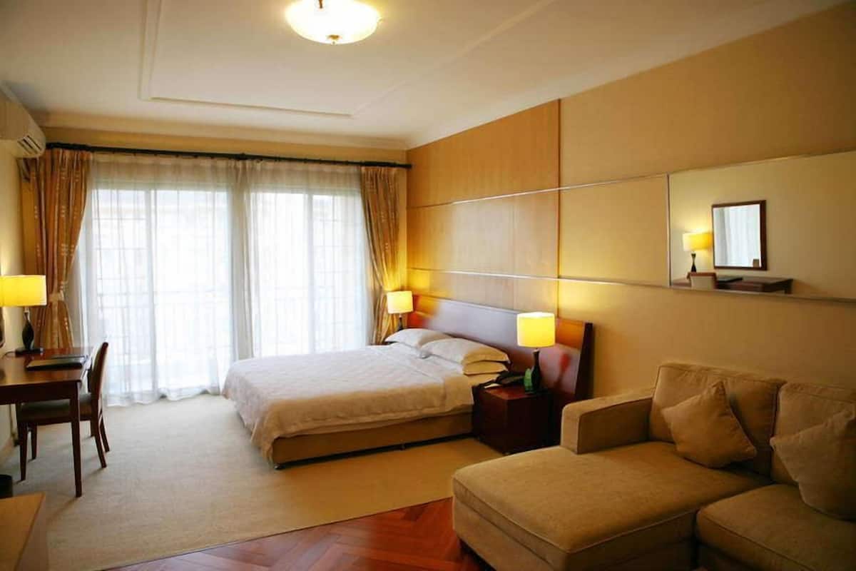 A hotel room with a bed, bedside tables, a desk, a chair, a sofa, and a large window with curtains.