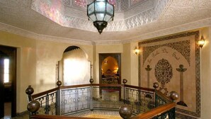 A grand staircase with ornate railings and a patterned floor, leading to a richly decorated room with a large, ornate wall tapestry and a hanging lantern. - Palais Dar Ouladna (Marrakech)