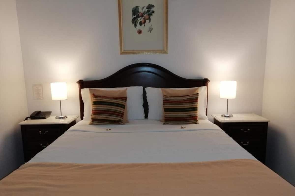 Una cama bien hecha con cabecera oscura, dos lámparas de noche y un cuadro en la pared.