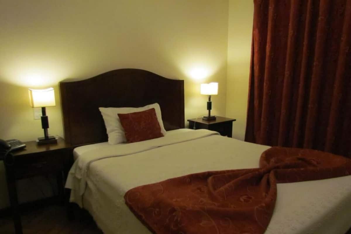 Una cama bien hecha con una colcha blanca y una manta marrón. Hay dos lámparas de noche a cada lado de la cama.