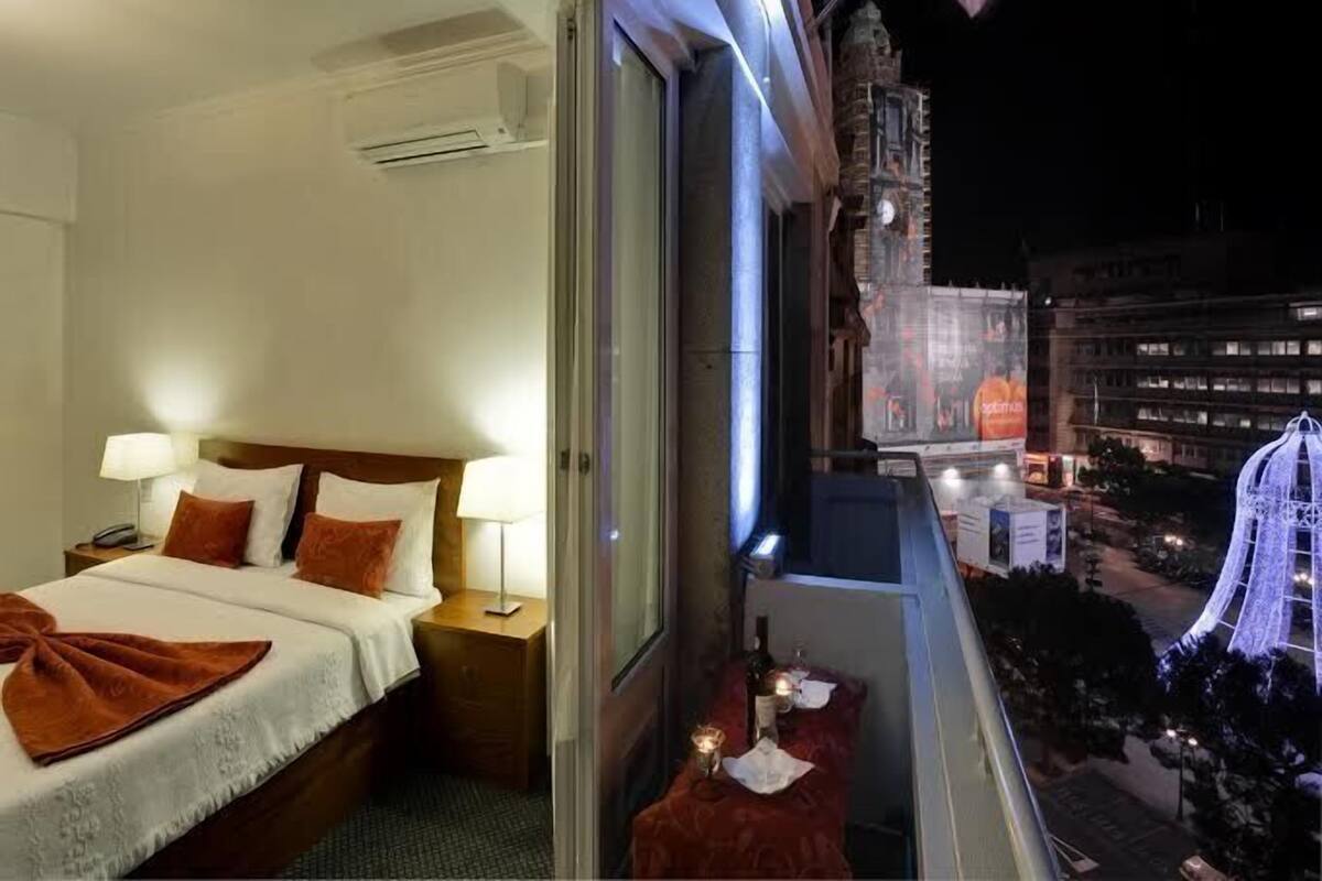 Habitación de hotel con cama, mesitas de noche y vista a un paisaje urbano nocturno.