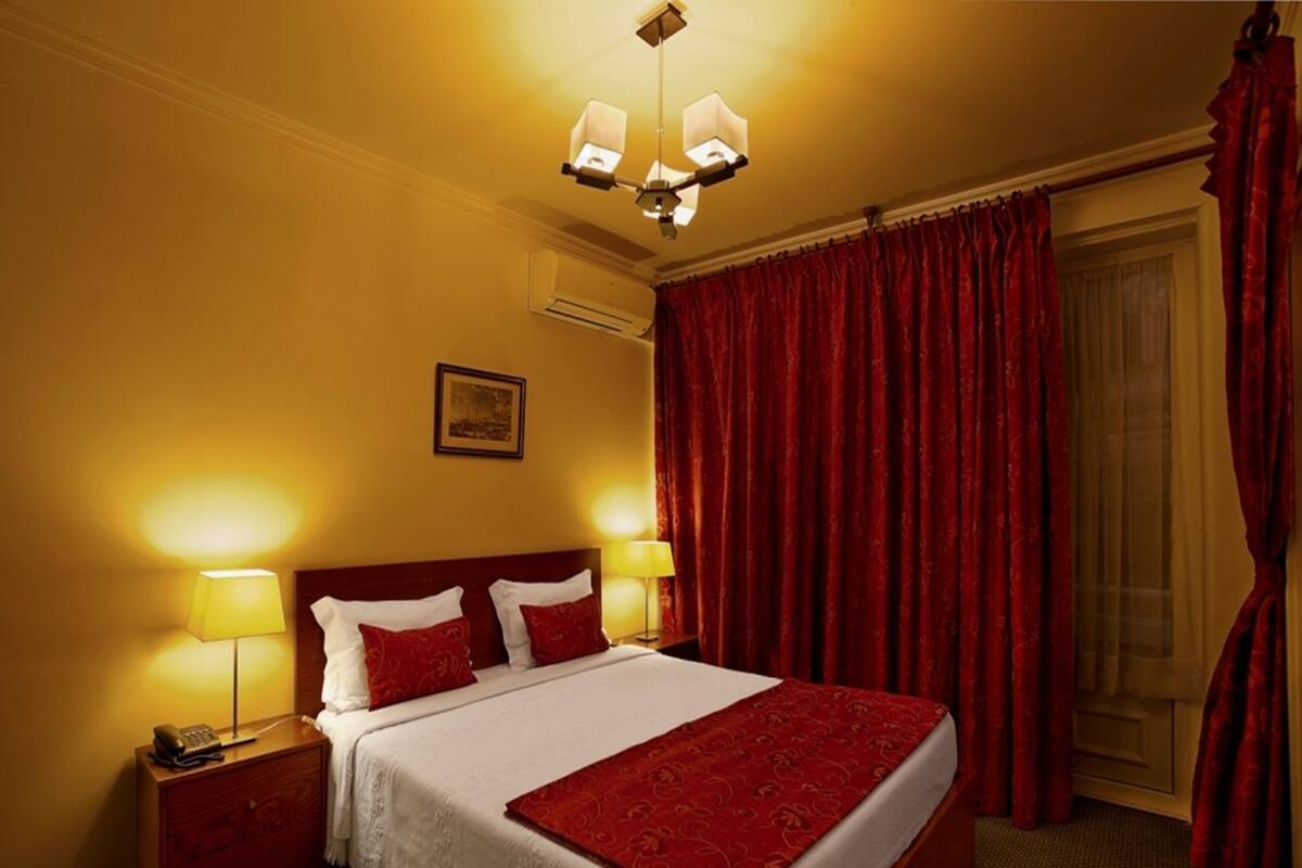 Una habitación de hotel con una cama grande, dos lámparas de noche, un teléfono, una mesita de noche, una cortina roja y un cuadro en la pared.