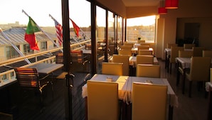 Un ristorante con tavoli e sedie disposti per consumare i pasti, ampie finestre con bandiere e vista sugli edifici circostanti.