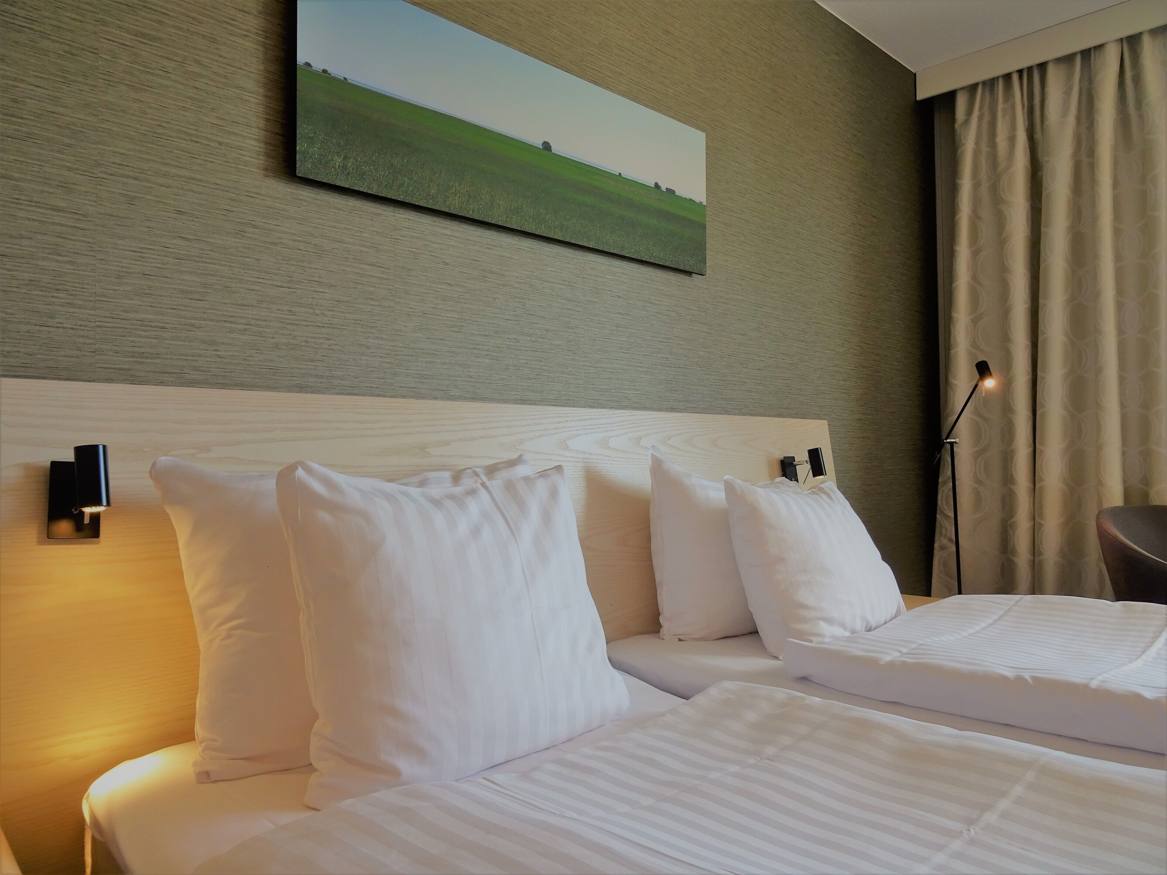A hotel room with two beds, a headboard, a painting on the wall, a lamp, and a curtain.