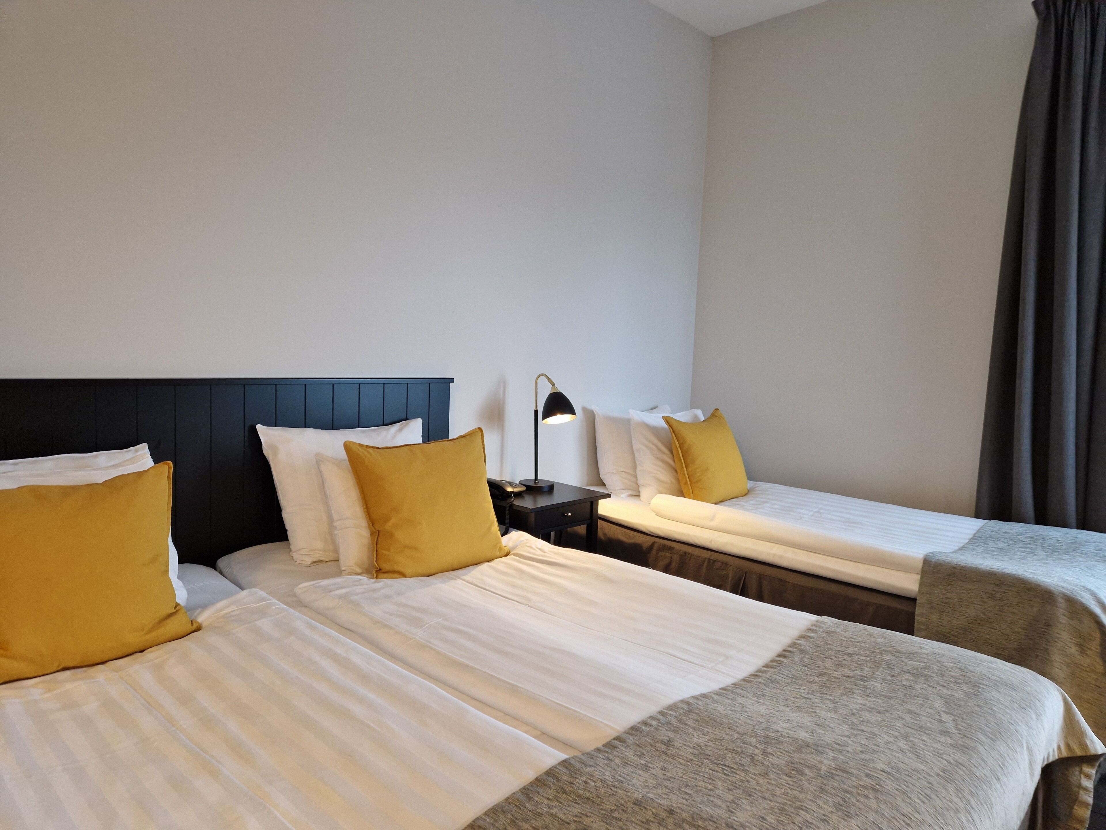 A hotel room with two beds, each with a headboard, a bedside table with a lamp, and a window with curtains.