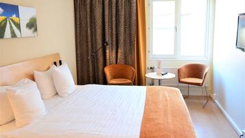 Een hotelkamer met een groot bed, twee stoelen, een tafeltje en een raam met gordijnen.